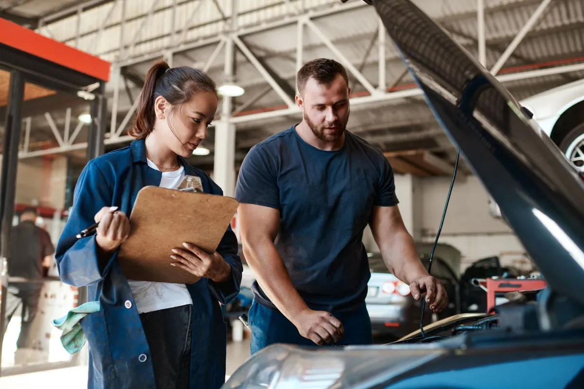 Two mechanics inspect the engine of a blue car in a busy auto repair shop, one taking notes on a clipboard while the other points at the engine hood.