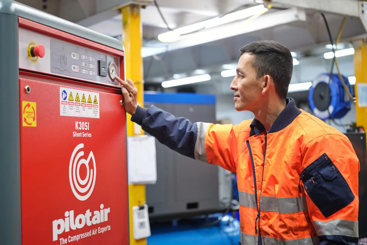Maintenance worker in an orange safety jacket presses controls on a red Pilotair machine in an industrial setting.
