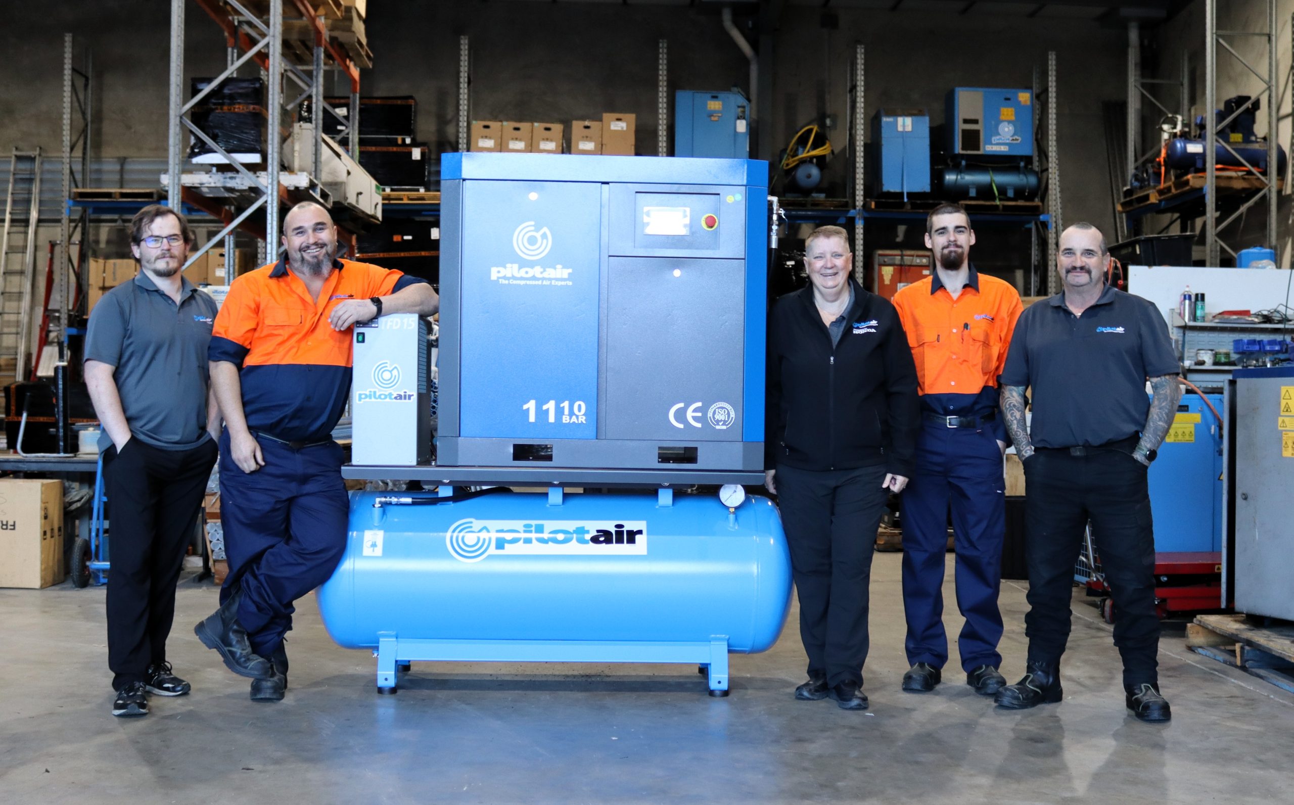 Six technicians pose in a workshop beside a large blue Pilot Air industrial compressor.