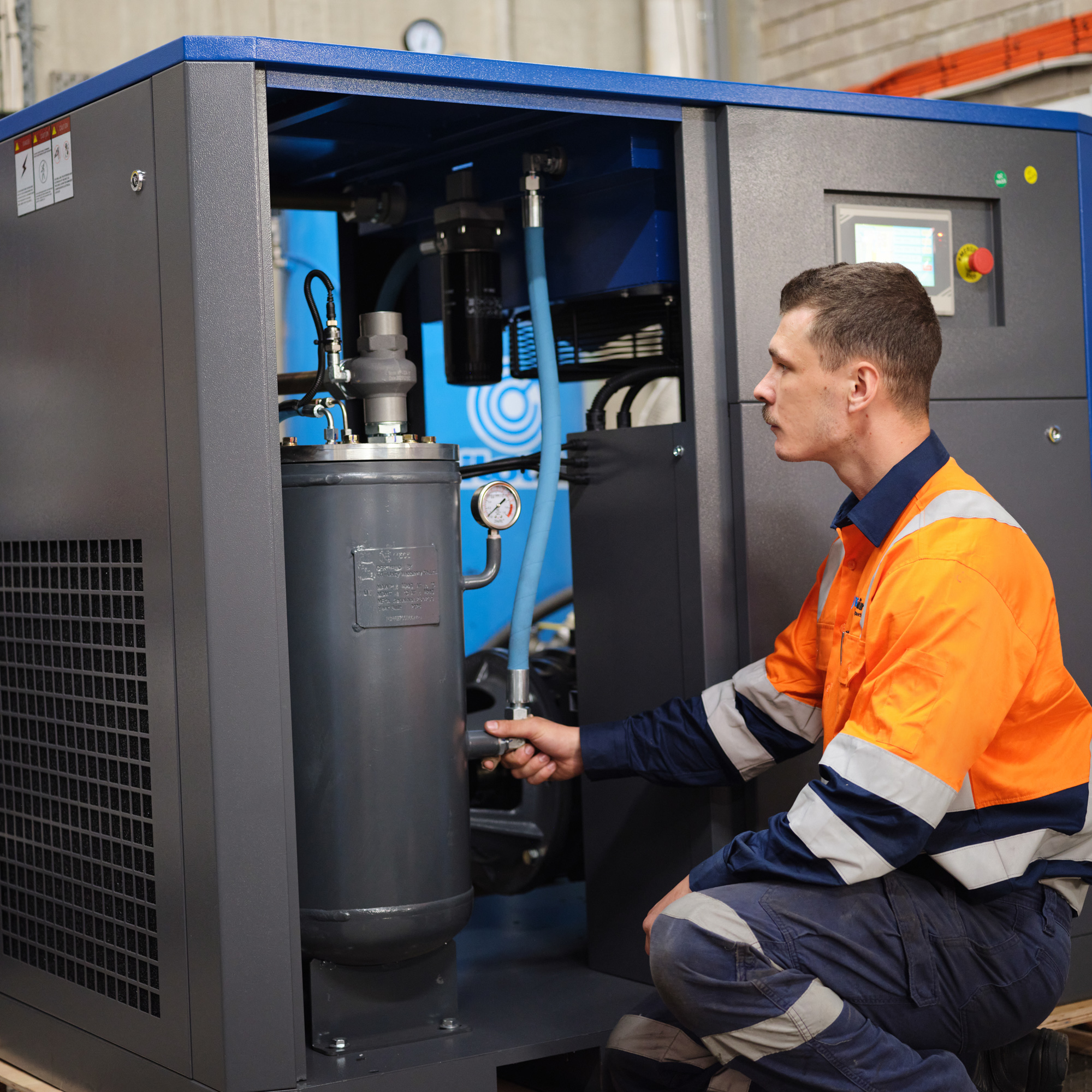 Maintenance worker in an orange high-visibility suit kneels beside a large gray industrial cabinet, inspecting pipes and gauges.